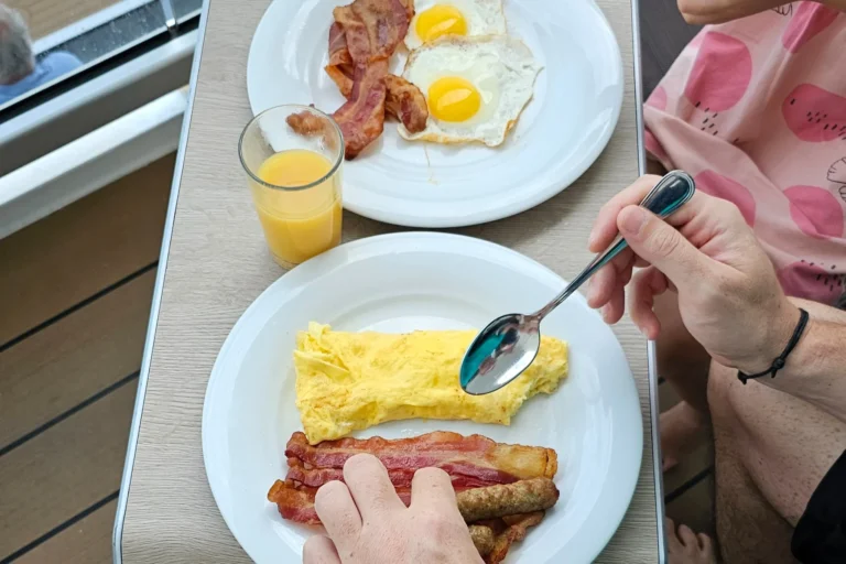 Frühstück auf dem Balkon einer Kreuzfahrtkabine mit Rührei, Spiegelei, Bacon und Orangensaft auf See.