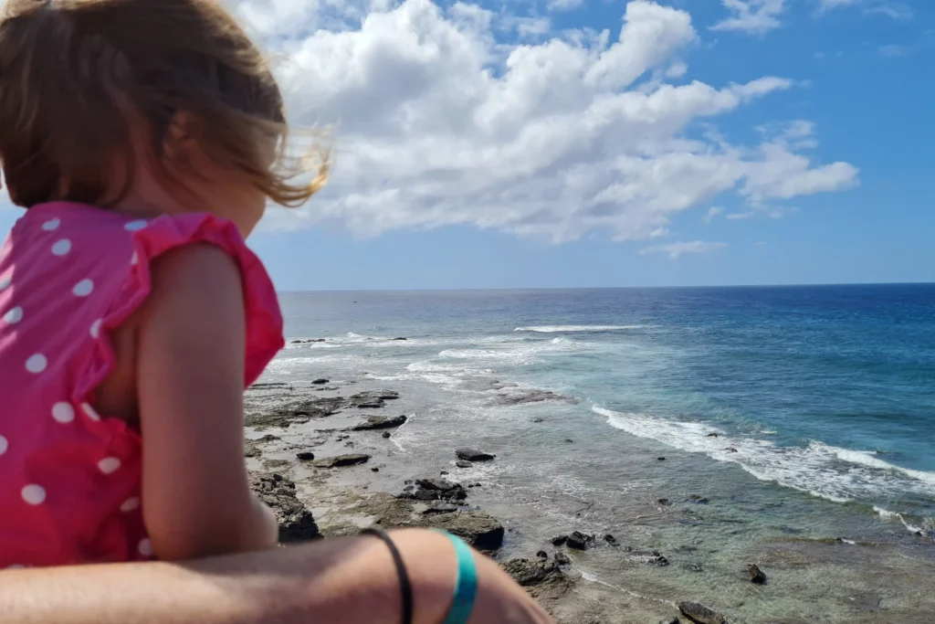 Kind blickt von der Küste auf das Meer auf Fuerteventura, felsige Küste mit Wellen und blauem Himmel.