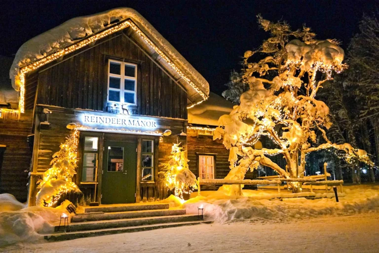Reindeer Manor mit Glasiglu in Levi, Lappland im Winter bei Nacht mit warm beleuchtetem Holzhaus und verschneiter Umgebung